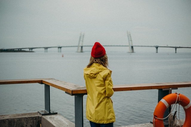 Woman in a yellow raincoat and a red hat standing next to the sea.