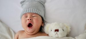 a baby yawning in bed next to a plushie toy
