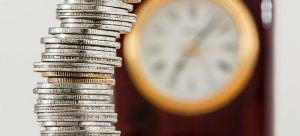 A clock in the background and pile of coins in front;
