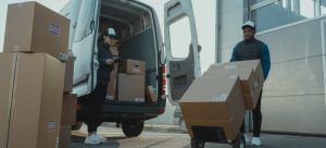A woman is carrying a phone while standing in front of the van full of boxes, a man is pushing a trolley with boxes loading on it;